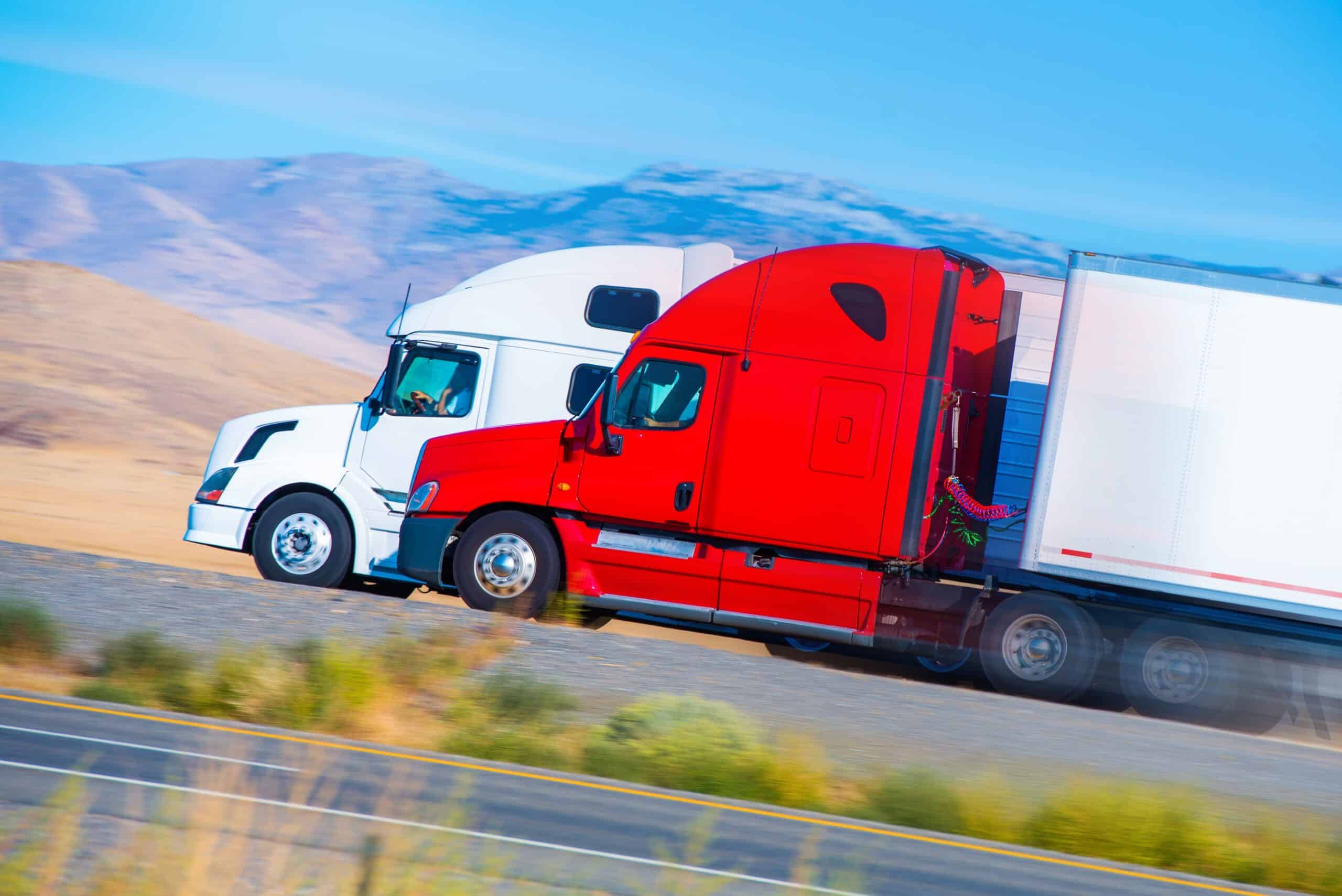 Two semi trucks next to each other driving up an incline with mountains in the background.