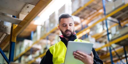 A focused warehouse worker in a high-visibility vest using a tablet to check inventory near the pallet storage racks.