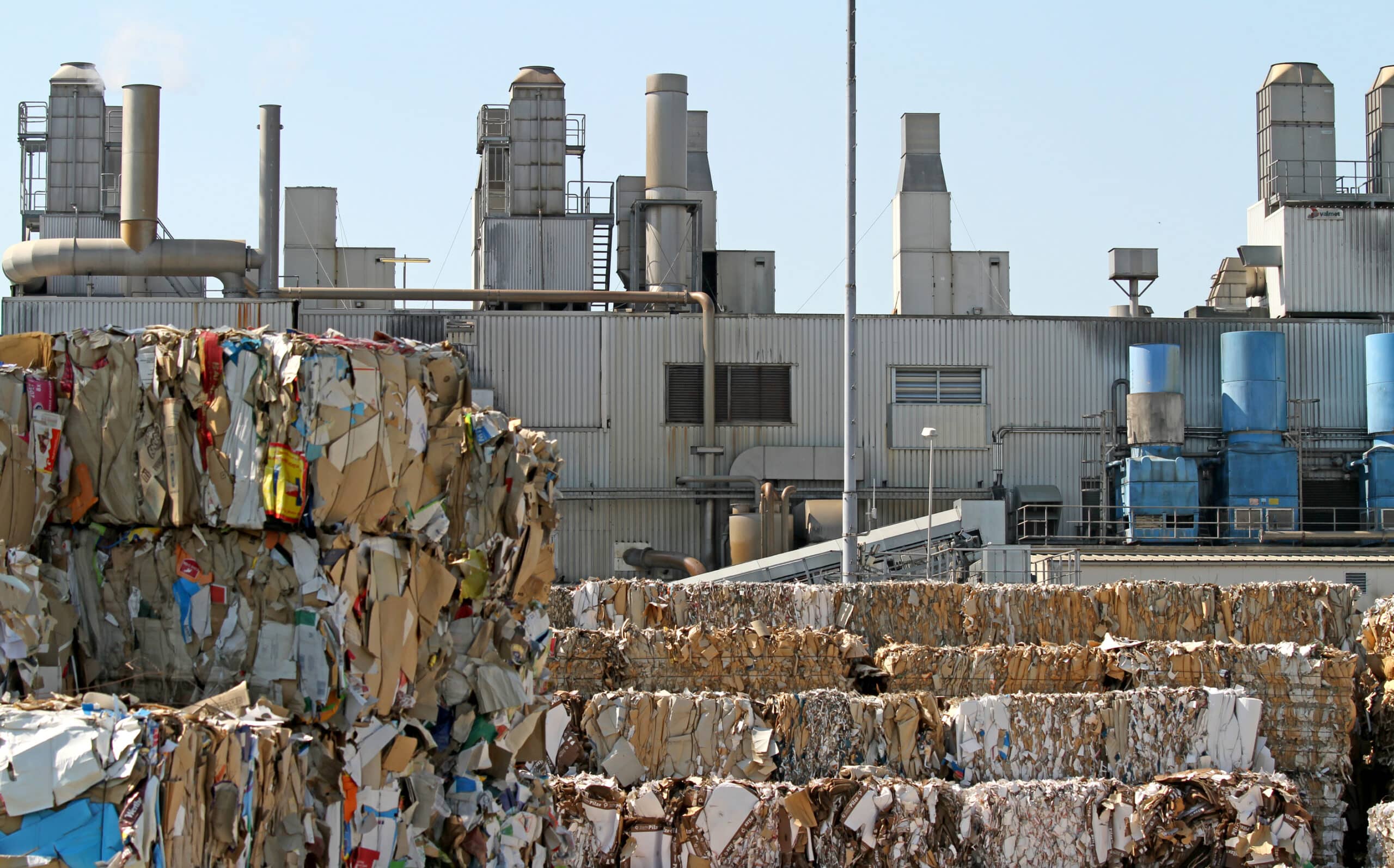 Stacks of compacted and bound recyclable materials in the foreground with an industrial recycling facility in the background, illustrating waste management and recycling operations.