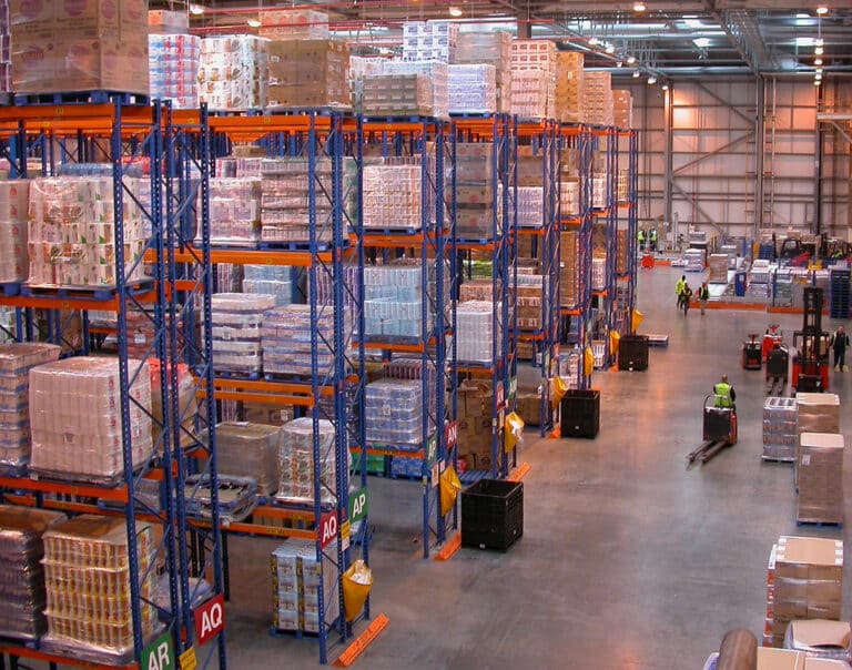 Interior of a warehouse with high pallet rack shelving filled with various goods and a worker operating a forklift.