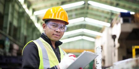 A focused industrial worker in a safety vest and hard hat holding a clipboard in a manufacturing plant.