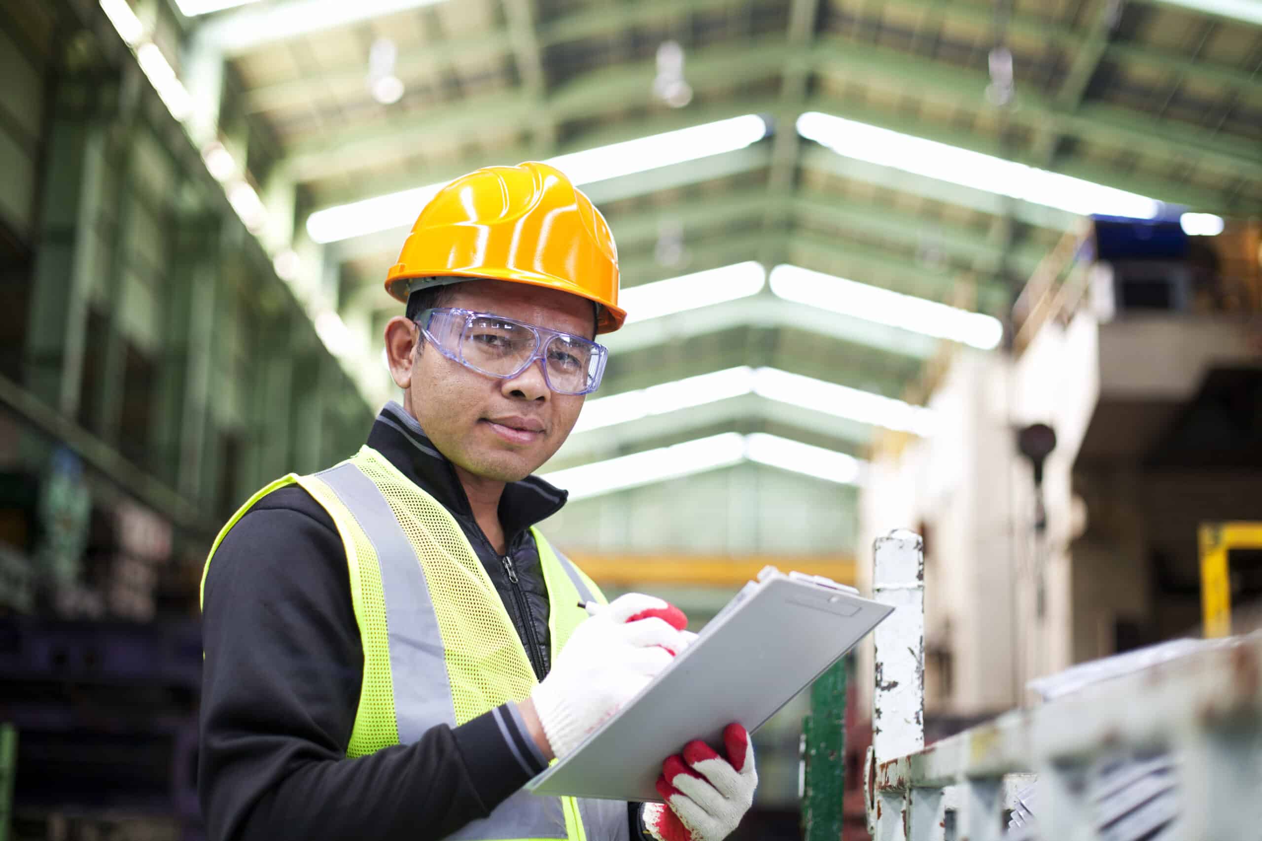 A focused industrial worker in a safety vest and hard hat holding a clipboard in a manufacturing plant.