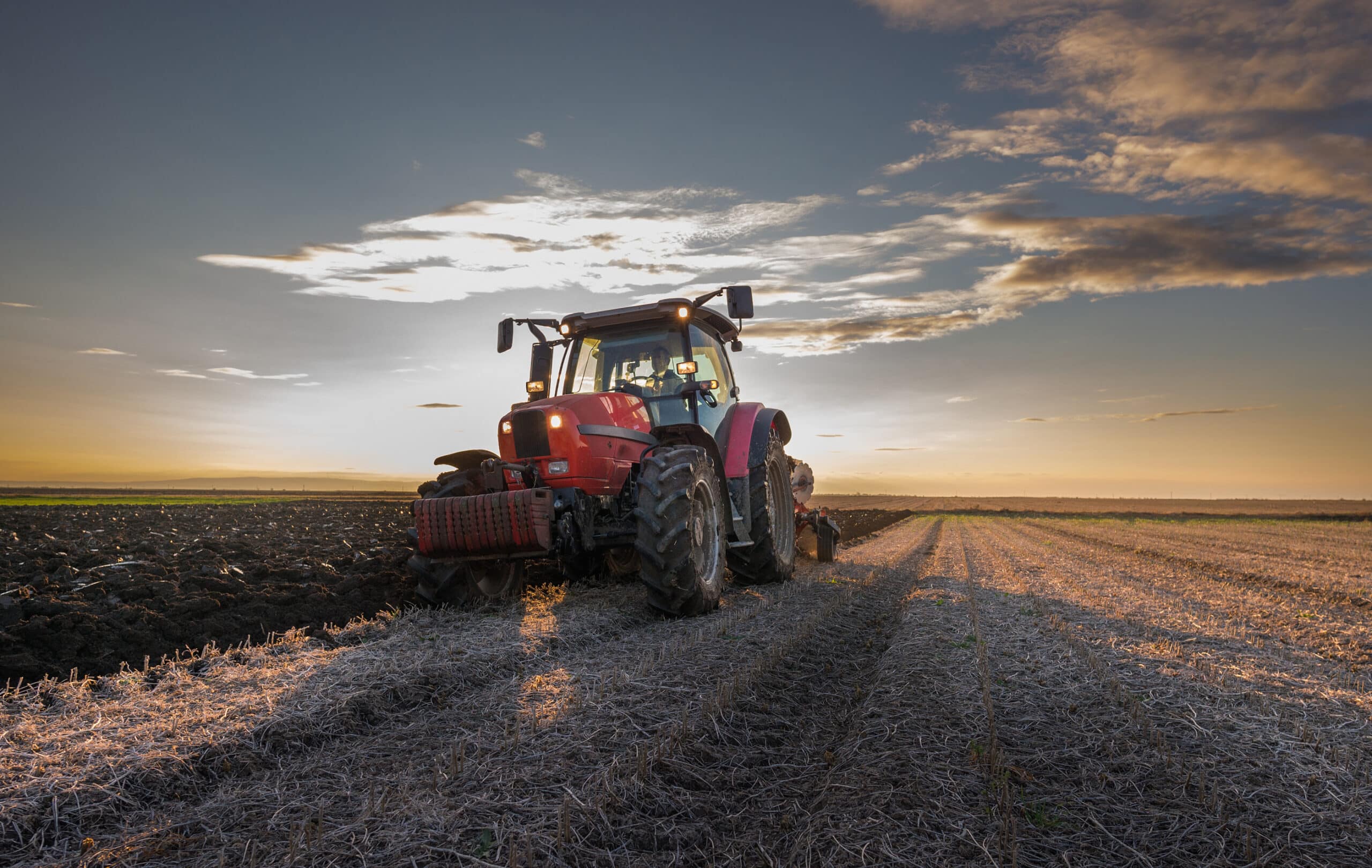 A tractor plowing the field at sunset, with rays of sunlight breaking through the clouds, casting a warm golden light over the freshly turned soil.