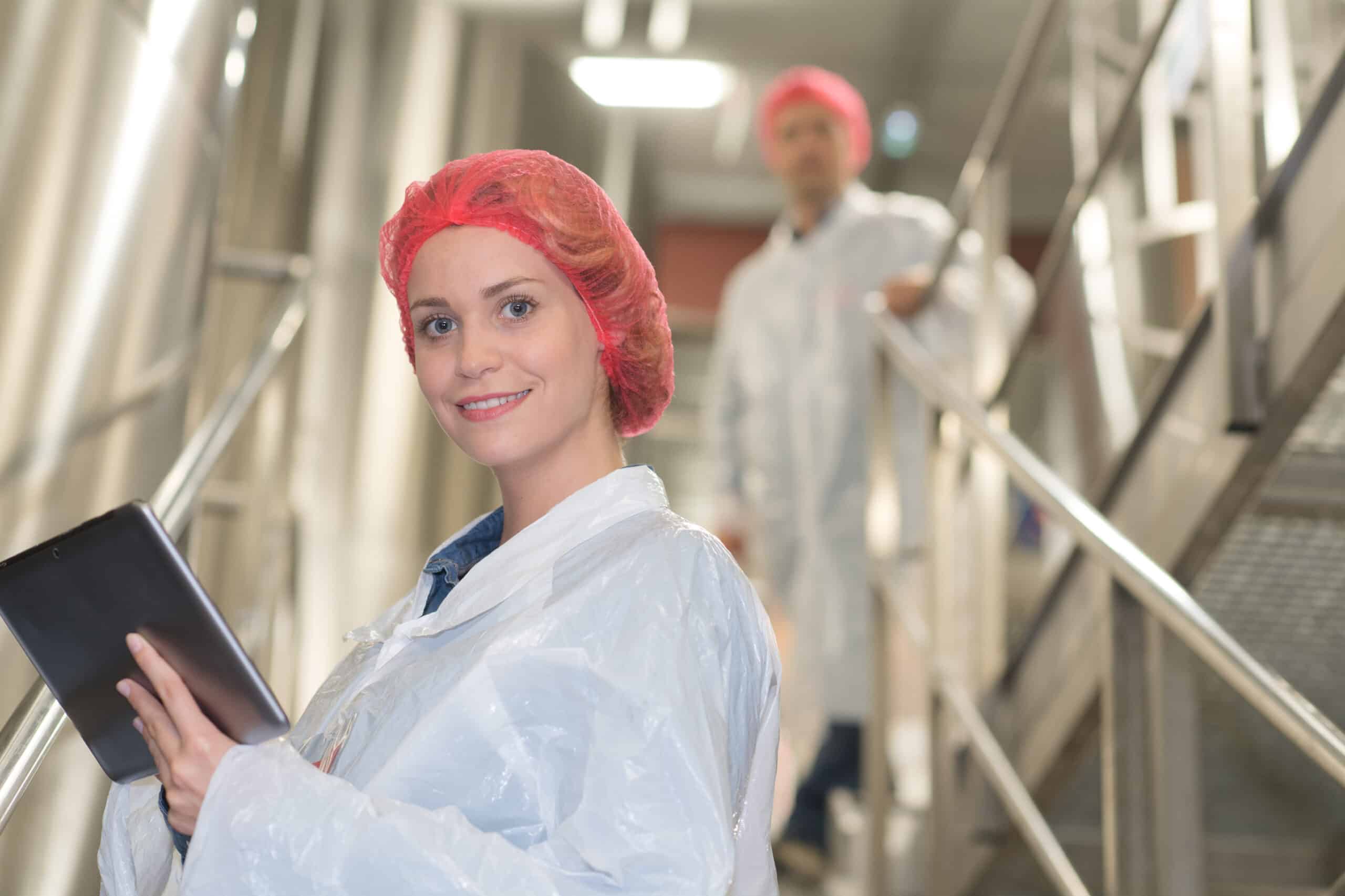 Quality control specialist with a tablet in a food processing facility, wearing protective clothing and hairnet, with a colleague in the background ensuring safety and hygiene standards.
