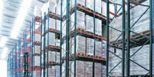 Bright warehouse with 11 visible rows of dark green racking filled with shrink-wrapped pallets and one red order picker transporting goods.