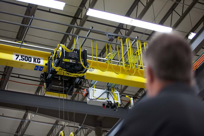 An industrial interior with a man observing a yellow overhead crane supporting advanced machinery, marked with a load capacity of 7,500 kg. with bright lighting overhead.