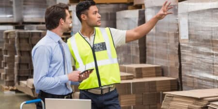 Two men in a warehouse; one in a suit and the other wearing a safety vest and pointing at something, with shelves of packaged products in background.