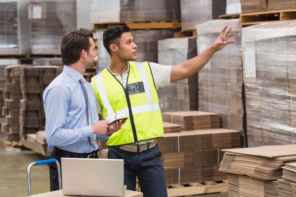 Two men in a warehouse; one in a suit and the other wearing a safety vest and pointing at something, with shelves of packaged products in background.