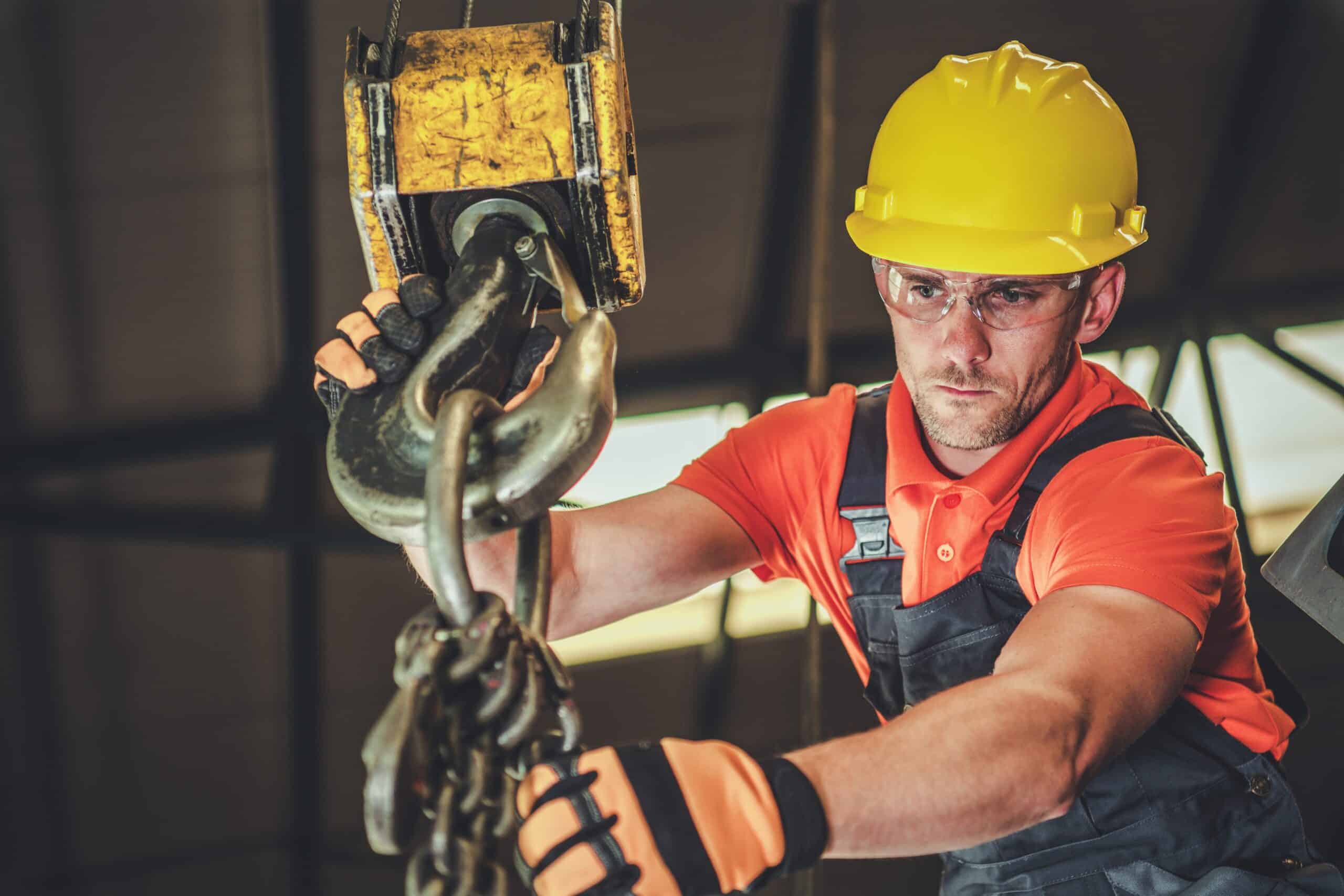 A technician wearing overalls and a hard hat up in the rafters of a warehouse inspecting chains hanging from a crane hook.