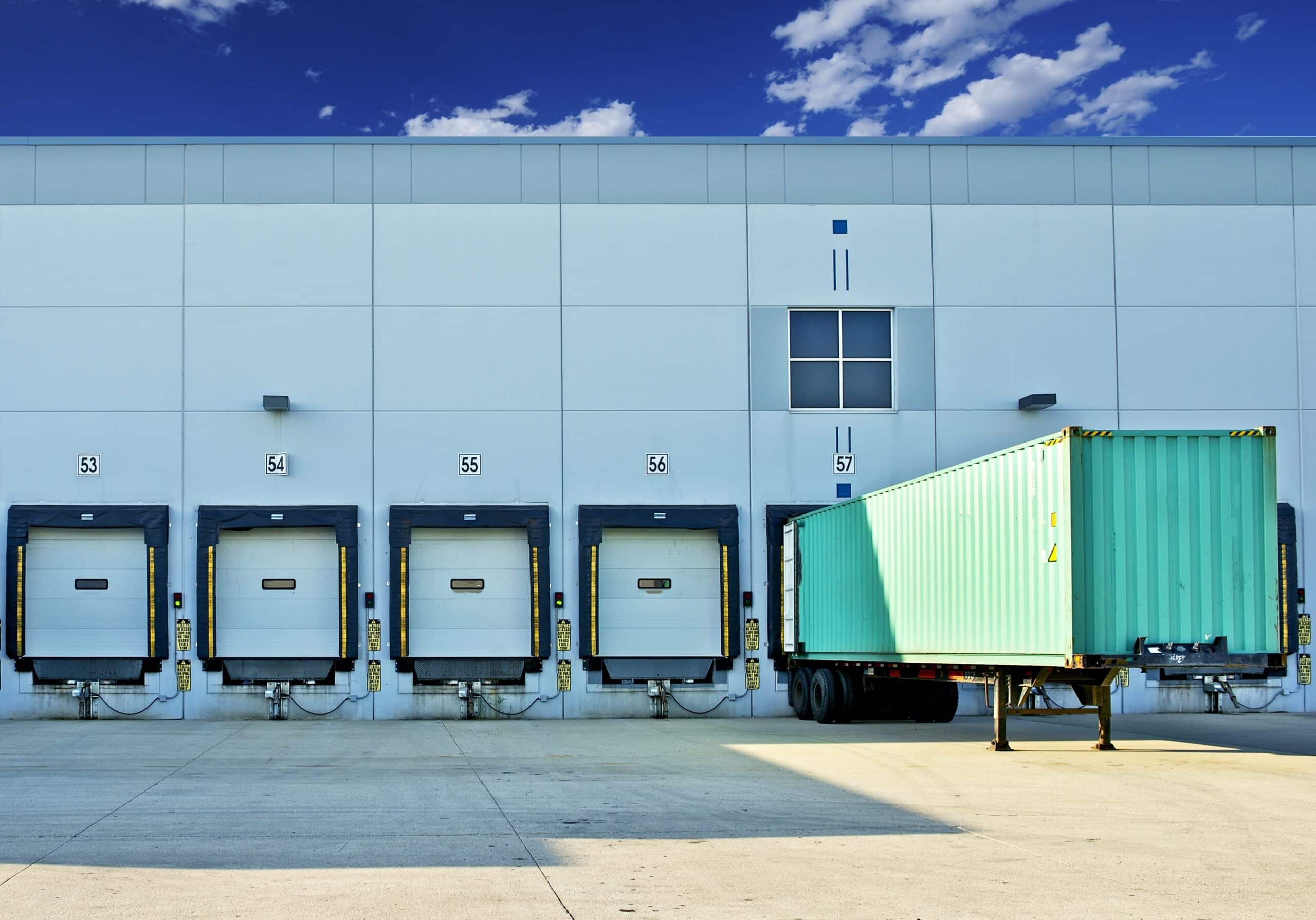 A blue sky above a distribution center with multiple loading docks. dock numbers 53 to 57 are visible, with a teal shipping container parked at dock 57.