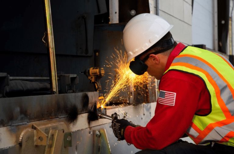 A worker in a reflective vest and helmet uses a power tool on metal, creating sparks, with a visible American flag on the sleeve.