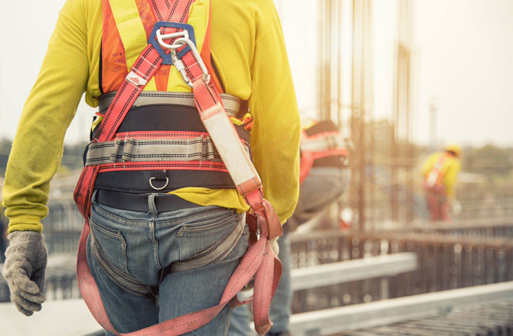 A construction worker wearing a safety harness and gloves stands on a site, with other workers visible in the background. focus on safety gear and construction environment.
