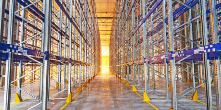 looking down a row of empty blue and steel racking in a warehouse with yellow sunlight streaming in at the very end of the row.