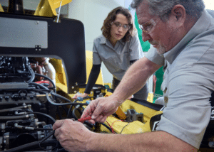 Two people work on machinery in a workshop. One person, wearing safety glasses, uses pliers on a wire, while the other observes attentively. Both are dressed in work shirts and the machinery appears to be a part of a large, yellow industrial vehicle.