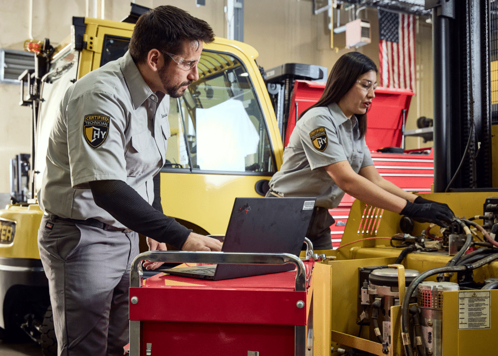 Two technicians communicating to each other while one is checking hydraulic lines on a industrial machine and the other is logging information on a laptop.