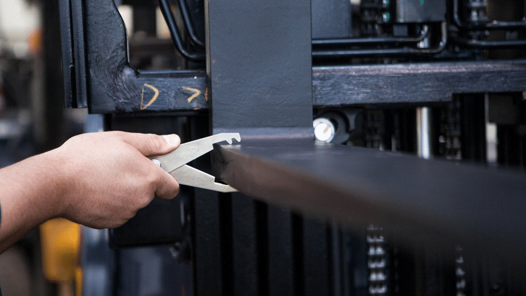 A close-up image of a hand using a measuring clamp tool to inspect a fork attached to a forklift.