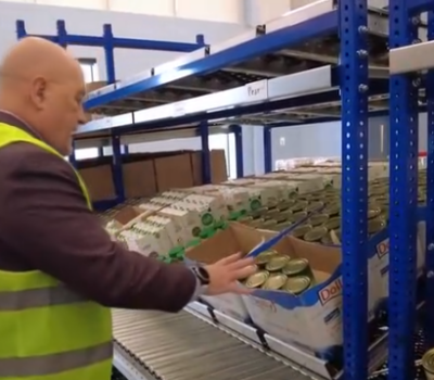 an image of a guy facing and explaining the new pushback racking system and how it will help volunteers. Viewer sees boxes filled with cans on the racking system in front of the man.